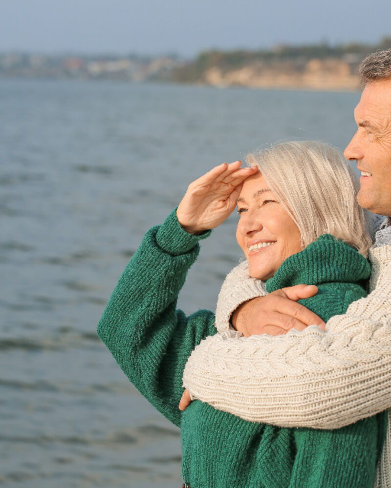 Senhora idosa a sorrir e a ser abraçada pelo marido junto ao mar